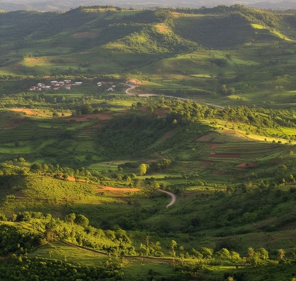 A scenic aerial view of lush green hills and valleys in Ghana, with terraced farmlands, winding roads, and a small village nestled in the landscape under soft, golden sunlight.